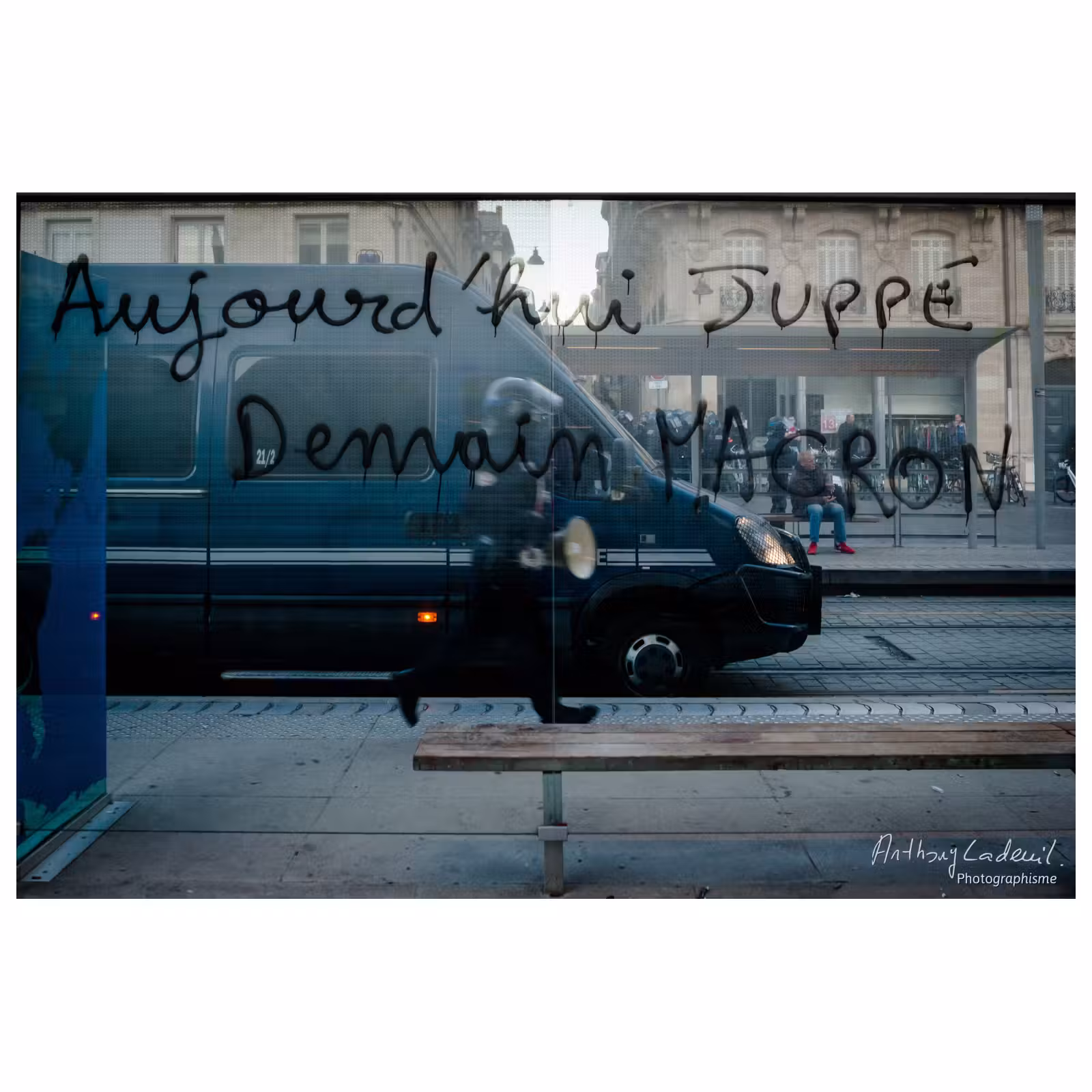 tags menaçants contre Emmanuel Macron sur les vitres d'un abri de bus avec un fourgon de CRS en arrière plan