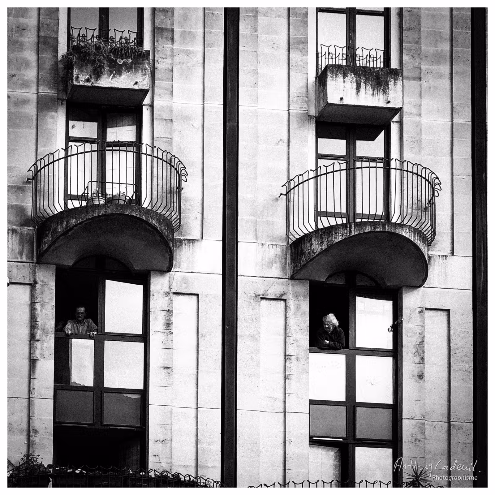 Photographie noir et blanc d'une façade d'immeuble avec deux personnes à leurs fenêtres