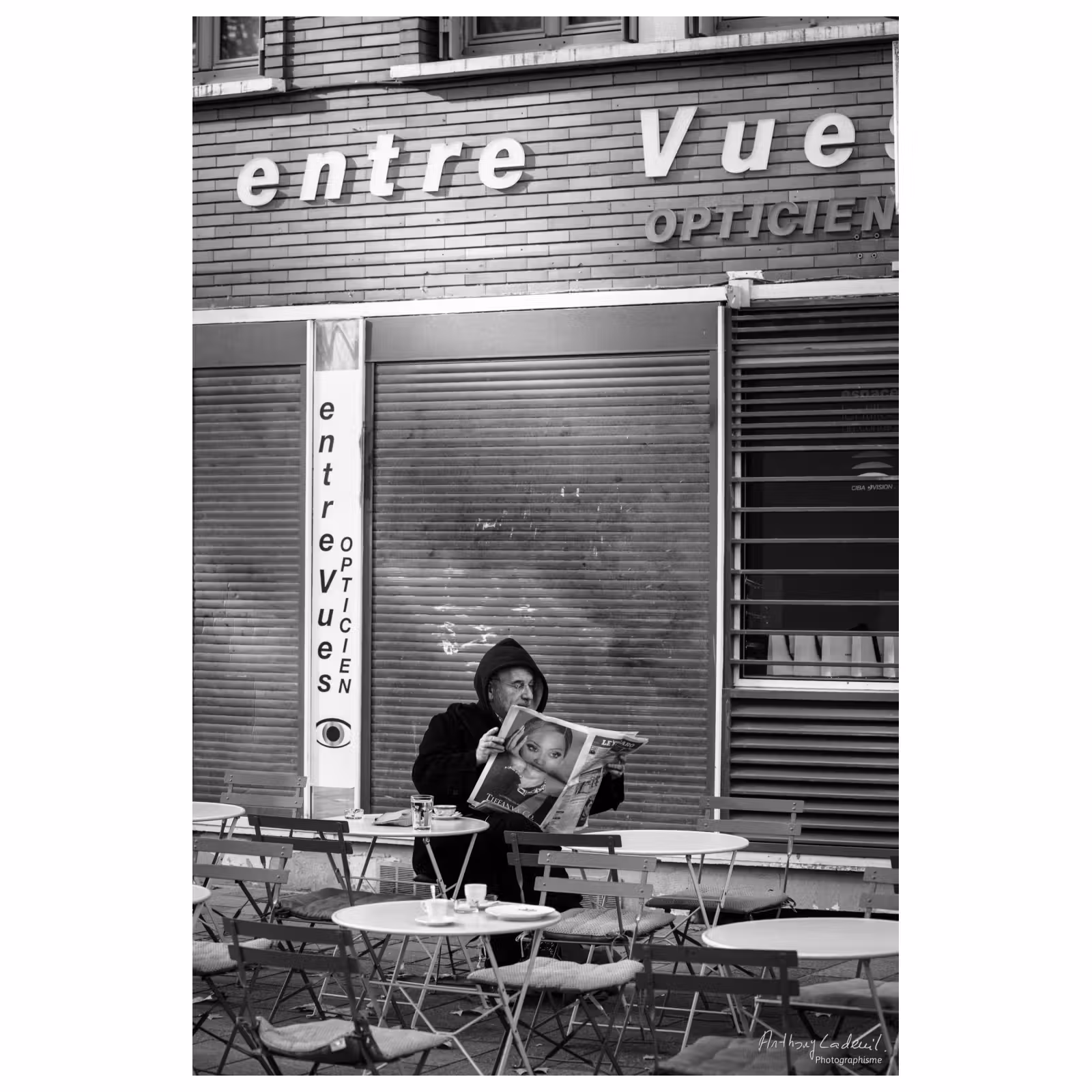 photographie d'un homme lisant le journal sur une terrasse devant un magasin d'optique